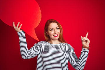 Sticker - Young beautiful redhead woman holding speech bubble over red isolated background very happy pointing with hand and finger to the side