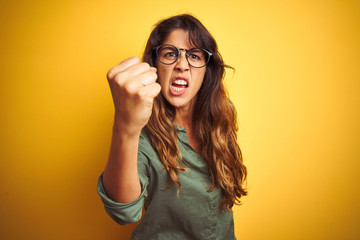 Wall Mural - Young beautiful woman wearing green shirt and glasses over yelllow isolated background angry and mad raising fist frustrated and furious while shouting with anger. Rage and aggressive concept.