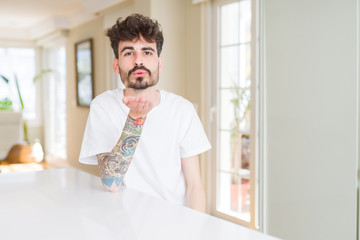 Poster - Young man wearing casual t-shirt sitting on white table looking at the camera blowing a kiss with hand on air being lovely and sexy. Love expression.