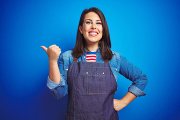 Young beautiful business woman wearing store uniform apron over blue isolated background smiling with happy face looking and pointing to the side with thumb up.