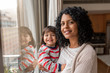© mavoimages - Smiling mother and daughter standing by their living room window