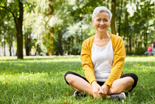 Positive Friendly Looking Caucasian Female Pensioner Sitting On Grass In Stylish Sportswear And Sneakers, Keeping Her Legs Crossed, Having Rest After Cardio Training On Warm September Day, Smiling