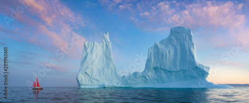Fotomural Little red sailboat cruising among floating icebergs in Disko Bay glacier during midnight sun season of polar summer