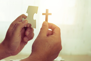 Wall Mural - close up of a man hands holding jigsaw and cross over jigsaws on wooden table against window light, christian concept show Jesus is most important part in human life with copy space