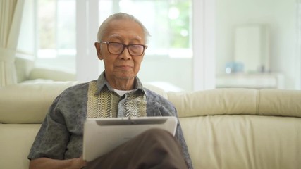 Wall Mural - Happy senior man using a digital tablet while sitting on the sofa in the living room at home. Shot in 4k resolution