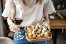 Girl Holding Glass Red Wine And Wooden Plate With Cheese. Delicious Cheese Mix With Walnuts, Honey. Tasting Dish On A Wooden Plate. Food For Wine.