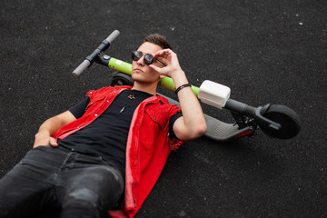 Sticker - Cool young hipster man in a stylish red vest with a trendy hairstyle in vintage jeans is lying and straightens fashionable sunglasses. Handsome guy resting after riding an electric scooter. Menswear.