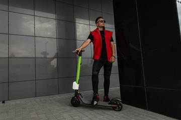 Sticker - American young hipster man with a hairstyle in stylish denim red-black clothes in sunglasses stands with a modern electric scooter near a dark wall in the city. Cool guy model is resting outdoors.