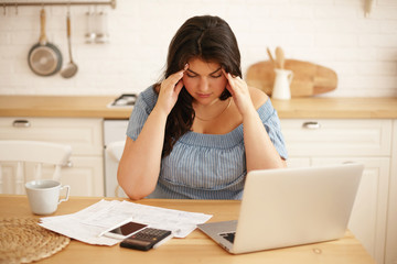 Stressful depressed young overweight woman facing financial problems, having frustrated look, holding hands on her face, sitting at kitchen table with laptop, doesn't have money to pay mortgage