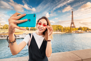 Wall Mural - Asian girl in a stylish outfit and pink glasses takes a selfie photo on the background of the main attractions of Paris - the Eiffel tower on the banks of the river Seine