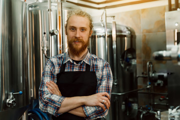 A portrait of handsome brewer with dreadlocks in uniform at the beer manufacture with metal containers on the background, who is making beer on his workplace in the brew-house.