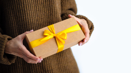 Woman in brown sweater holding gift box with yellow ribbon in hands on white background