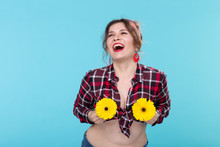 Portrait Of Positive Young Woman In Plaid Shirt Holding Two Yellow Flowers With Different Hearts Near Breast Posing Against Blue Background. Concept Of Body Beauty In All Forms, Body Positivity