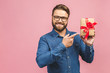 Happy holiday, my congredulations! Portrait of an attractive casual man giving present box and looking at camera isolated over pink background.