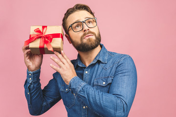 Happy holiday, my congredulations! Portrait of an attractive casual man giving present box and looking at camera isolated over pink background.