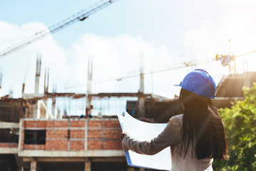 Wall Mural - Asian woman architect wearing blue safety helmet checking working progress at contruction site.