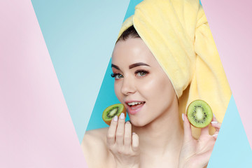 Portrait of young beautiful woman with healthy glow perfect smooth skin holds two piece of kiwi near face. Model with natural make up, with towel on head look into the hole of pink, blue paper.