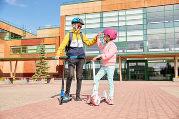 Poster - education, childhood and people concept - happy school children in helmets with backpacks and scooters making high five outdoors