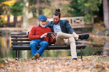 Senior Father And His Son Sitting On Bench In Nature, Using Tablet.