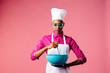 Portrait of a young woman in cooking hat and apron mixing a bowl with a whisker, isolated on pink studio background