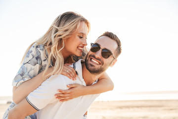 Wall Mural - Photo of unshaven happy man smiling and giving piggyback ride seductive woman while walking on sunny beach