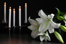 White Lilies And Blurred Burning Candles On Table In Darkness, Closeup With Space For Text. Funeral Symbol