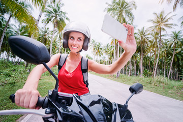 Wall Mural - Traveling lifestyle and technology. Outdoor portrait of pretty young woman in helmet sitting on scooter taking selfie on her smart phone.