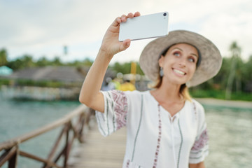 Wall Mural - Vacation and technology. Young woman taking selfie with her  smartphone on tropical beach. Focus on cellphone.