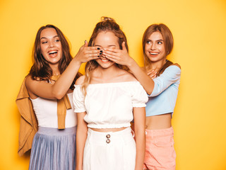 Wall Mural - Three young beautiful smiling hipster girls in trendy summer clothes.Sexy carefree women posing near yellow wall in studio.Models surprising their friend.They cover her eyes and hugging from behind