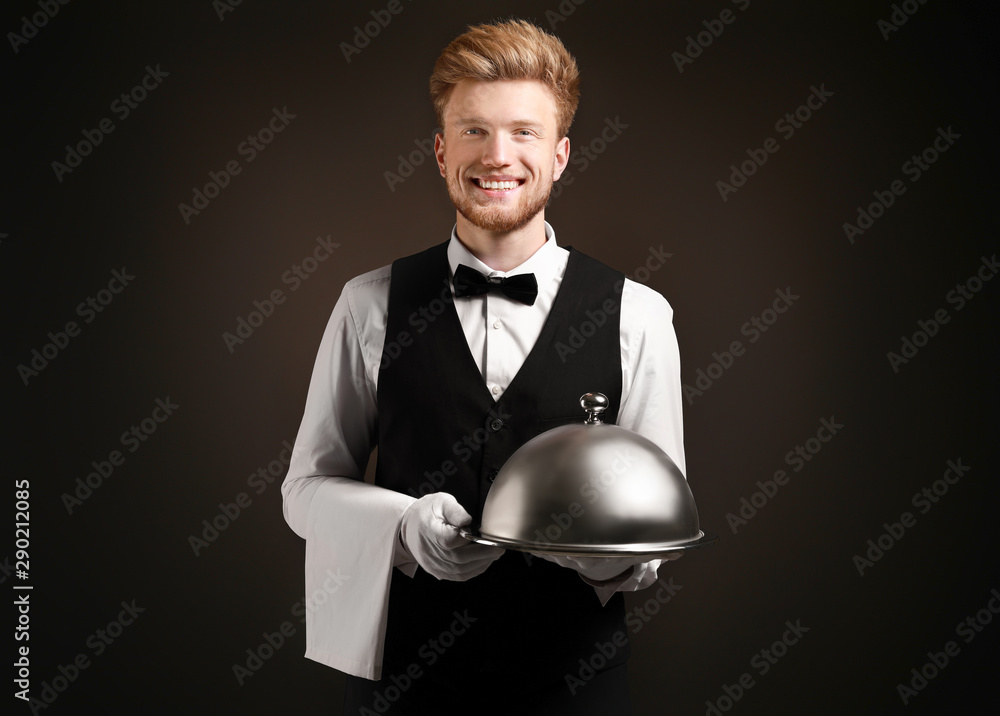 Handsome waiter with tray and cloche on dark background