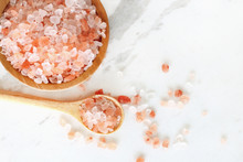 Top View Of Himalayan Pink Rock Salt In Wooden Bowl And Spoon On White Marble Table.
