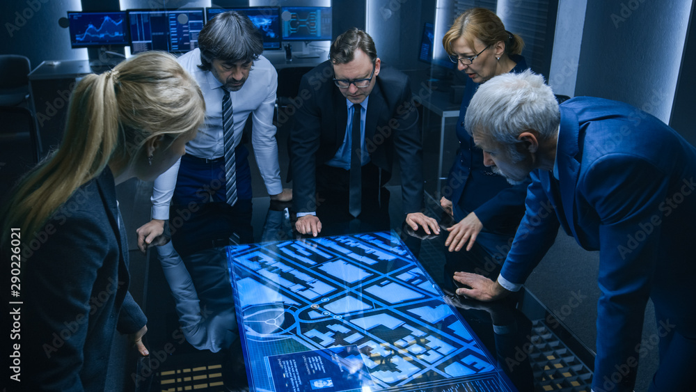 High Angle Shot of Team of Government Intelligence Agents Talking while Standing Around Digital Touch Screen Table and Tracking Suspect. Satellite Surveillance Operation in the Monitoring Room. - obrazy, fototapety, plakaty 