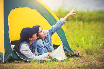 Wall Mural - Two close friend Asian friendship relax in camping tent in green meadow on lake side view background. Girl pointing finger to sky. People lifestyle travel on vacation concept. Summer picnic activity