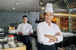 © DragonImages - Portrait of confident Asian chef standing with arms crossed and looking at camera with young cook preparing food in the background in the kitchen