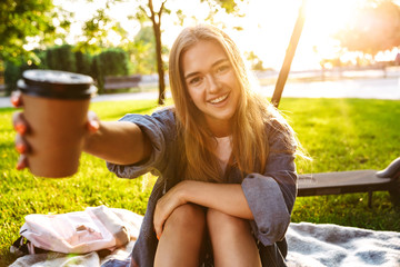 Wall Mural - Teenage girl sit on grass in nature green park showing cup of coffee.