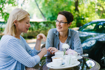 Wall Mural - Beautiful mature woman is talking to female friend in street cafe smiling having fun enjoying warm summer day. Modern lifestyle, friendship and conversation concept.