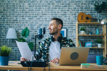 Attractive guy is recording podcast using microphone holding piece of paper indoors creating content for audio blog. People, occupation and sound equipment concept.