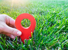 A Hand Is Holding A Red Location Marker In The Green Grass. The Concept Of Tourism And Travel. Navigation And Exploration. Destination. Holiday, Vacation. Buying Building Land. Selective Focus