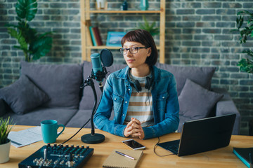 Portrait of young adult woman recording podcast for audio blog using mic, headphones and laptop working at table at home. Blogging, equipment and apartment concept.