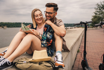 Wall Mural - Young couple on vacation having fun driving electric scooter through the city, they sitting on riverbank taking a break from driving.