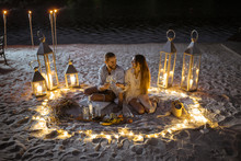 Couple Having A Romantic Dinner, Hugging And Clinking Wine Glasses At The Beautifully Decorated Place Illuminated With Different Lights The Sandy Beach At Dusk
