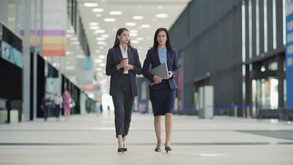 Wall Mural - Full length wide shot of two businesswomen walking down office lobby at break and having talk. Young woman holding takeaway coffee cup, middle aged colleague carrying laptop computer