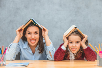 Cheerful mom and daughter on a gray background. During this, on the head of my mother and daughter there are open books. Looking at the camera show the tongue.