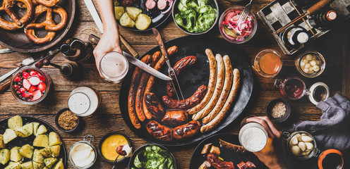 Flat-lay of Octoberfest dinner table with grilled sausages, pretzel pastry, potatoes, cucumber salad, sauces, beers and people drinking and eating over wooden background, top view, wide composition