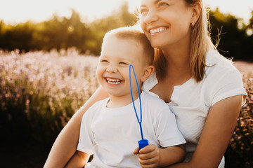 Wall Mural - Lovely young mother playing with her son outdoor. Sweet little boy laughing while doing soap bubbles with his mother against sunset.