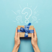 Female Hands Hold A Gift Box With A Blue Ribbon On A Blue Background With A Question Mark. Surprise Concept, Waiting For A Gift For The Holidays, Birthday, Christmas, Wedding. Flat Lay, Top View