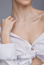 Cropped Shot Of A Lady, Wearing White Chest Tied Shirt. She Has Silver Necklace With Suspension In A Shape Of Hexagon On Her Neck. Her Right Hand Is Touching Shoulder. 