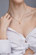 Cropped shot of a lady, wearing white chest tied shirt. She has golden chain with a pendant on her neck. Her right hand is touching shoulder. The photo is taken by grey background. 