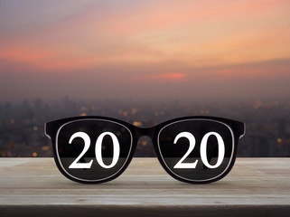 Wall Mural - 2020 white text with black eye glasses on wooden table over blur of cityscape on warm light sundown, Business vision happy new year 2020 concept