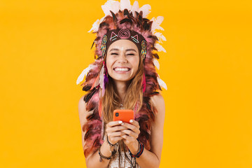 Sticker - Positive young emotional indian woman in carnival suit isolated over yellow wall background using mobile phone.